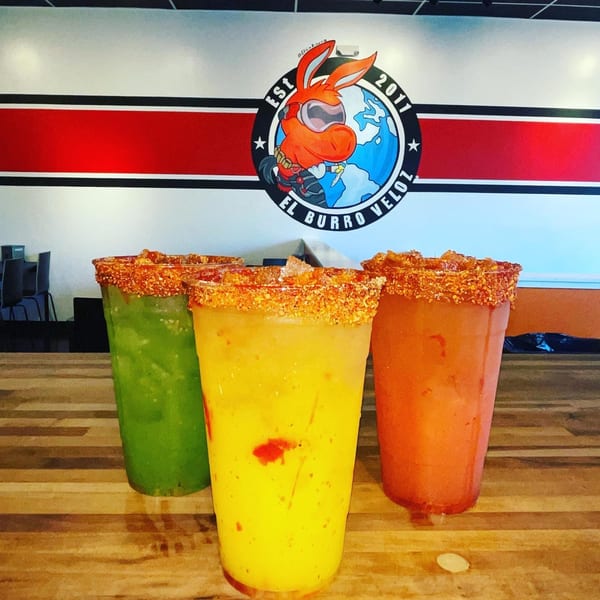 Three colorful drinks with chili-lime rims on a wooden table, with the El Burro Veloz logo in the background.