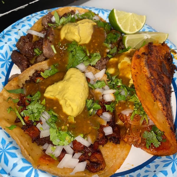 A plate of three tacos with various fillings, cilantro, onions, salsa, and lime wedges.