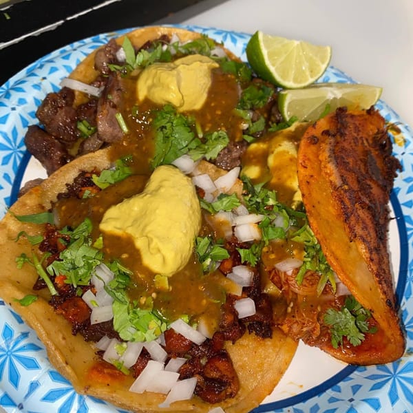 Three tacos with various fillings, salsa, cilantro, onions, and lime wedges on a paper plate.
