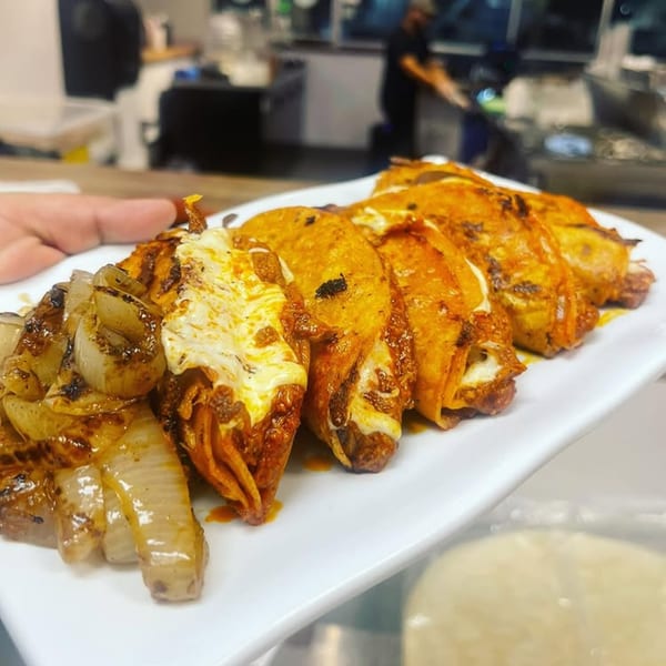 A plate of cheesy birria tacos with grilled onions, with a blurred kitchen in the background.